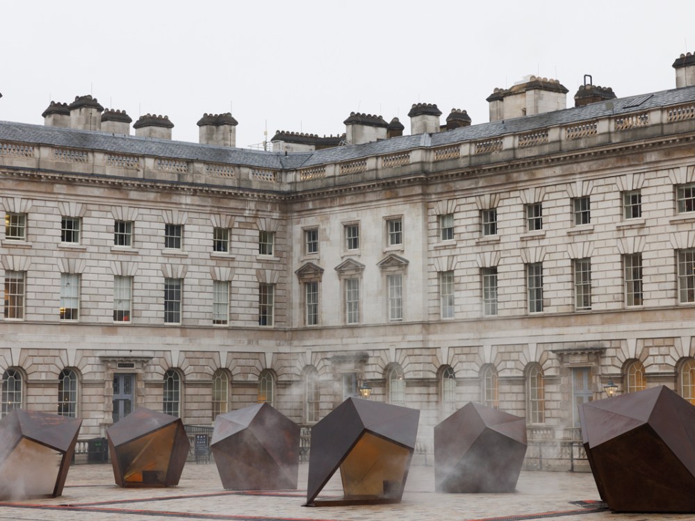 Several 3D, trapezoid-like, metal sculptures are installed in a courtyard in front of a Georgian-era building in London. Fog seems to rise from the sculptures.
